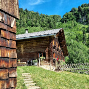 Dieser Walser Paarhof steht vor einem sattgrünen Wald und im Hintergrund leuchtet der blaue Himmel. Das alte Bauernhaus wird mit prächtigen Blumen geschmückt und davor gedeiht ein üppiger Bauerngarten. der von einem seltenen Zaun begrenzt wird.
